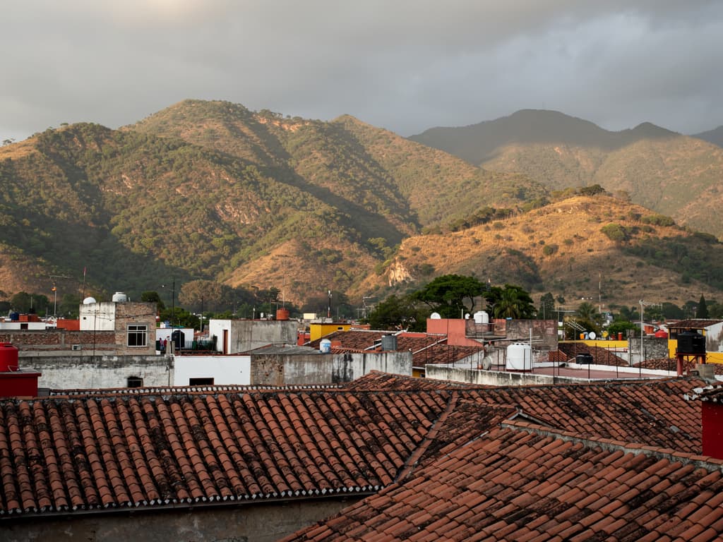Late afternoon from a rooftop in Oaxaca de Juárez. Terracotta roofs close in. Dry green-brown forested Sierra Madre hills and low ridges, no snow, no glaciers,