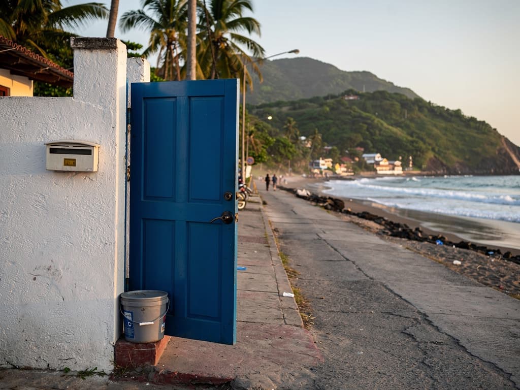 Blue door on a Zicatela side street, a plastic bucket holding the door open, late afternoon.